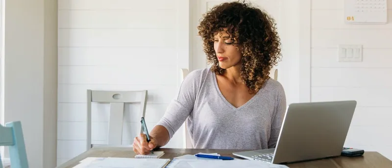 A woman with curly brown hair sits at a wooden table in a brightly lit room, writing in a small notebook while looking at papers spread out next to a silver laptop.
